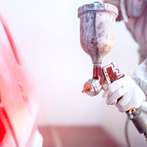 A man in a white suit uses spray painting services of Precision Powder Coating to apply a fresh, even red coat of paint to a car