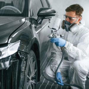 A man in a white suit uses spray painting services of Precision Powder Coating to apply a fresh, even black coat of paint to a car